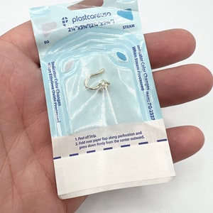 Puede incluir: Una bolsa de pl&aacute;stico blanca con texto azul y una tira indicadora azul y blanca. El texto en la bolsa dice "plastcareusa 2 1/4"x3 3/4" (2 1/2"x2 1/4") STEAM Indicator Color Changes item: PG-2337 When Steam Processed". La bolsa contiene un peque&ntilde;o aro de nariz dorado.