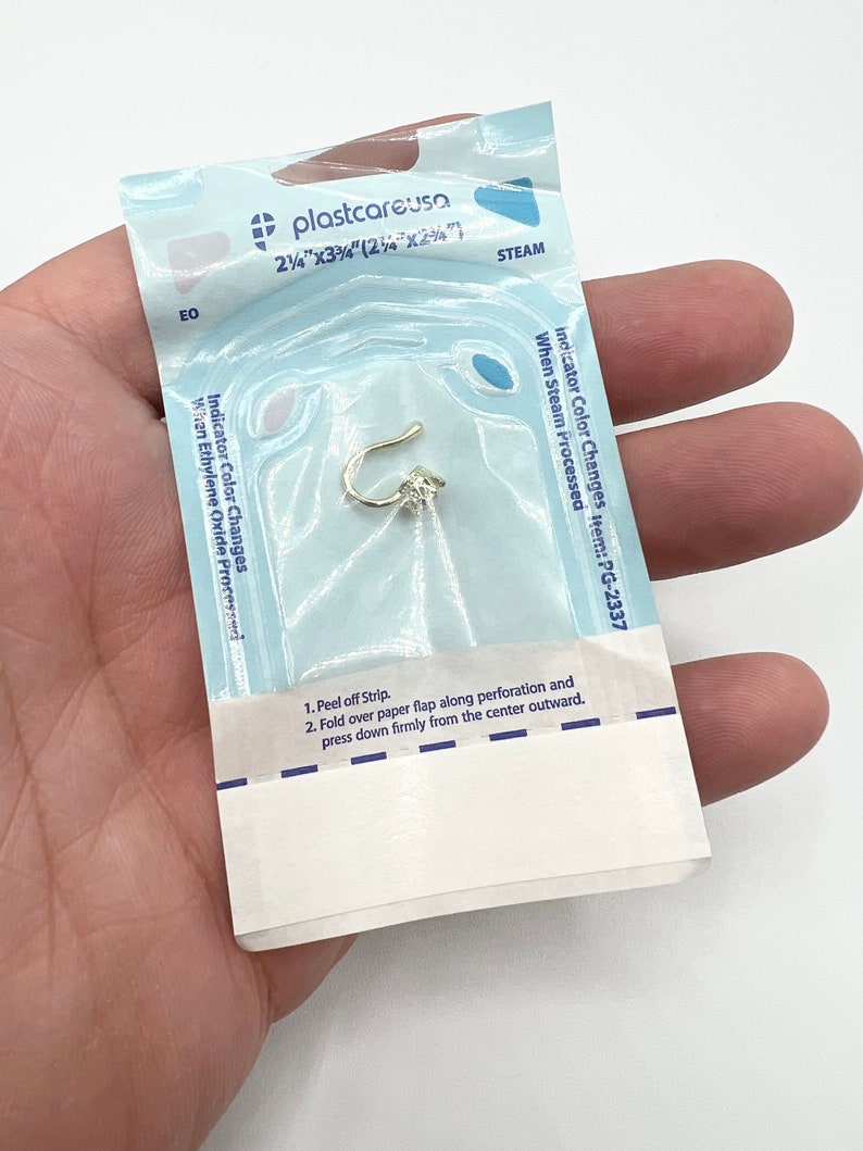 Puede incluir: Una bolsa de pl&aacute;stico blanca con texto azul y una tira indicadora azul y blanca. El texto en la bolsa dice "plastcareusa 2 1/4"x3 3/4" (2 1/2"x2 1/4") STEAM Indicator Color Changes item: PG-2337 When Steam Processed". La bolsa contiene un peque&ntilde;o aro de nariz dorado.