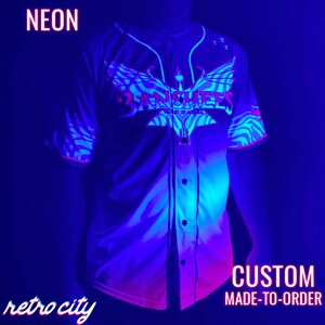 May include: A white baseball jersey with a pink and blue zebra print design. The jersey features a large pink and red graphic of a bird with the text "3ANSHERS" and "CITY OF ANGELS". The text "NEON" appears in the top left corner of the image and the text "CUSTOM MADE-TO-ORDER" appears in the bottom right corner of the image.