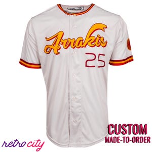 Puede incluir: Camiseta de béisbol blanca con la palabra "Arrakis" en escritura amarilla y naranja, y el número "25" en rojo. La camiseta tiene rayas rojas y amarillas en las mangas y el cuello. El texto "retro city" está en la parte inferior izquierda y "CUSTOM MADE-TO-ORDER" en la parte inferior derecha.