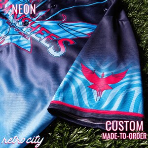 May include: A black and blue baseball jersey with a red and white design. The jersey has the words "NEON" and "ANCHEES" on the front. The sleeve has a red bird design. The text "CUSTOM MADE-TO-ORDER" is on the bottom of the image. The text "retro city" is on the bottom left of the image.