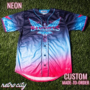 May include: A custom-made baseball jersey with a gradient design in black, blue, and pink. The jersey features a large graphic of a winged creature with the text "Banshees" and "City of Angels" on the front. The jersey is made to order and has a retro city logo on the bottom.