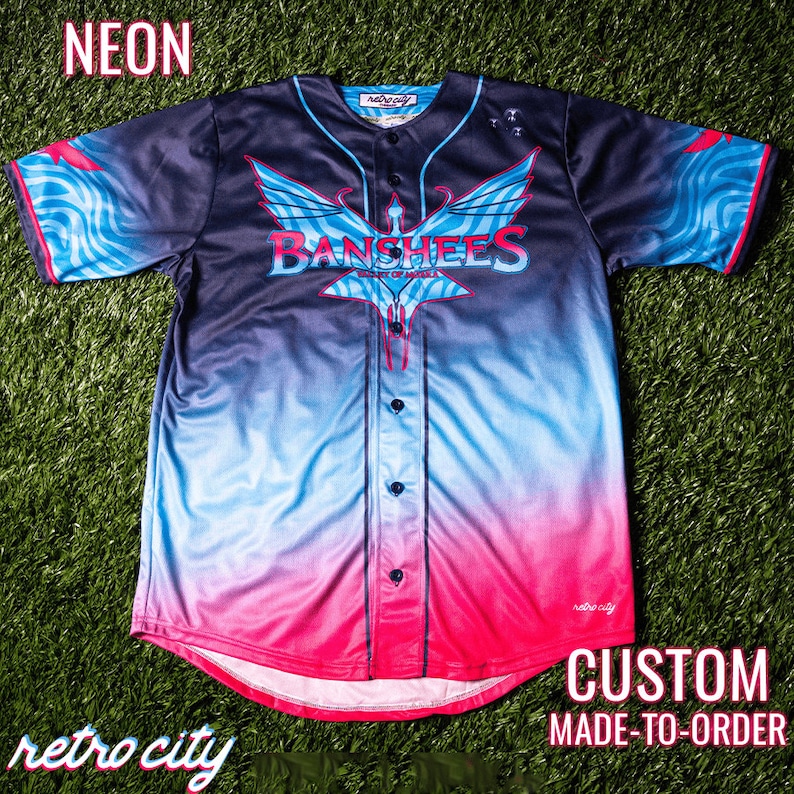 May include: A custom-made baseball jersey with a gradient design in black, blue, and pink. The jersey features a large graphic of a winged creature with the text "Banshees" and "City of Angels" on the front. The jersey is made to order and has a retro city logo on the bottom.