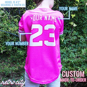 Op de afbeelding: Roze baseballshirt voor jeugd met witte letters. De achterkant van het shirt heeft "YOUR NAME" en het nummer "23" erop gedrukt. De tekst "CUSTOM MADE-TO-ORDER" is onder het shirt gedrukt.