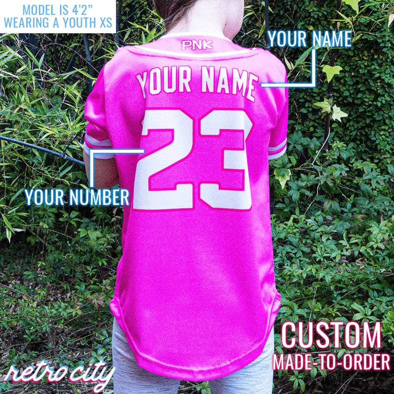 Op de afbeelding: Roze baseballshirt voor jeugd met witte letters. De achterkant van het shirt heeft "YOUR NAME" en het nummer "23" erop gedrukt. De tekst "CUSTOM MADE-TO-ORDER" is onder het shirt gedrukt.