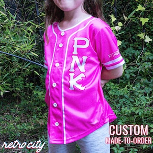 Peut inclure: Un maillot de baseball rose avec des bordures blanches et le mot "PINK" en grandes lettres blanches sur le devant. Le maillot est fait sur commande.