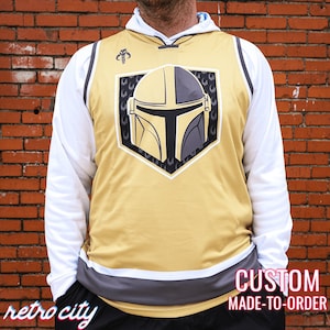 May include: Gold and gray jersey with a Mandalorian helmet graphic. The jersey has a white long-sleeve shirt underneath. The jersey has the words "retro city" and "custom made-to-order" printed on it.
