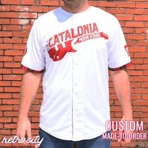 Puede incluir: Camiseta de béisbol blanca con texto rojo "CATALONIA MOON STARS" y gráfico de guitarra. Presenta una parte delantera abotonada, ribete a cuadros en las mangas y los textos "retro city" y "CUSTOM MADE-TO-ORDER".