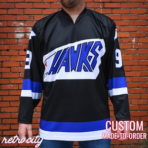 Op de afbeelding: Een zwart hockeytrui met witte en blauwe bies. De trui heeft een grote witte patch op de voorkant met het woord "HAWKS" in blauwe letters. Het nummer "9" staat op de rechter schouder van de trui.
