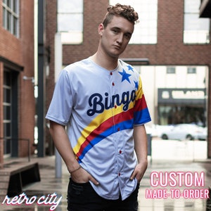 May include: A white baseball jersey with a blue, red and yellow stripe design. The word "Bingo" is written in a blue cursive font on the front of the jersey. There are three white stars on the right shoulder of the jersey. The jersey is custom made to order.