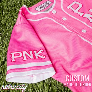 Op de afbeelding: Een roze baseballshirt met witte bies en het woord "PNK" in witte letters op de mouw. Het shirt heeft een knoopsluiting en is op bestelling gemaakt.