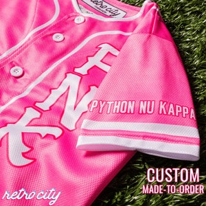 Op de afbeelding: Roze baseballshirt met witte letters die "Python Nu Kappa" op de mouw en "retro city" onderaan zeggen. Het shirt heeft een witte knoopsluiting en witte piping.
