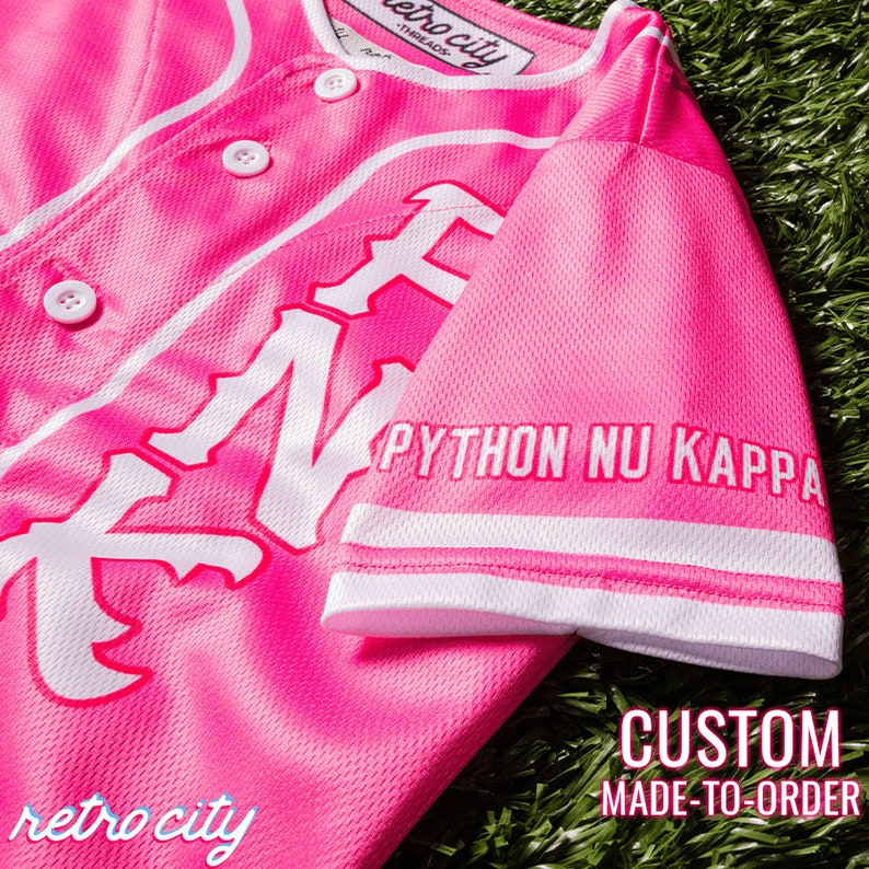 Op de afbeelding: Roze baseballshirt met witte letters die "Python Nu Kappa" op de mouw en "retro city" onderaan zeggen. Het shirt heeft een witte knoopsluiting en witte piping.