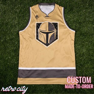 May include: A gold and black sleeveless jersey with a Mandalorian helmet graphic. The jersey has a white and gray stripe at the bottom and a small logo on the upper left. The text "retro city" is at the bottom left and "CUSTOM MADE-TO-ORDER" is at the bottom right.
