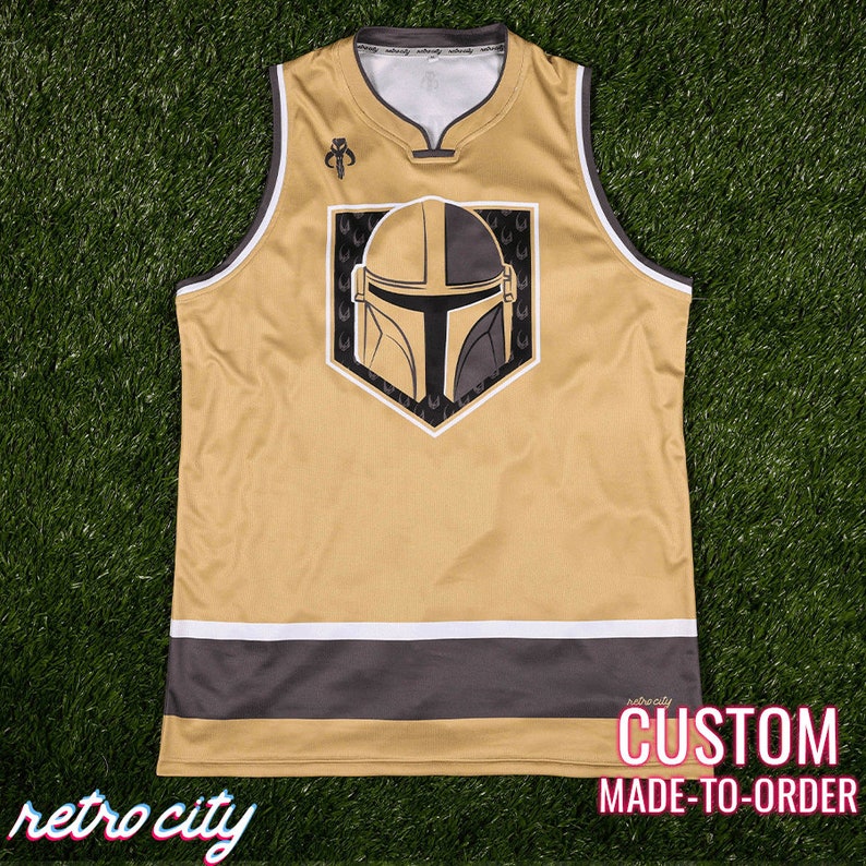 May include: A gold and black sleeveless jersey with a Mandalorian helmet graphic. The jersey has a white and gray stripe at the bottom and a small logo on the upper left. The text "retro city" is at the bottom left and "CUSTOM MADE-TO-ORDER" is at the bottom right.
