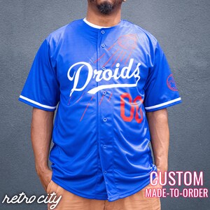 May include: A royal blue baseball jersey with white trim and red accents. The jersey features the word "Droids" in white cursive lettering and the number "00" in red. The jersey is custom made to order.