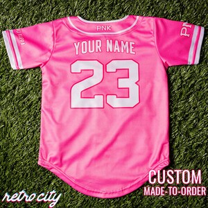 Op de afbeelding: Roze baseballshirt met witte bies en de tekst "YOUR NAME" en het nummer "23" op de rug. Het shirt is op maat gemaakt.