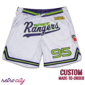 Op de afbeelding: Witte basketbalshorts met groene en paarse bies. De shorts hebben de tekst "Space Rangers" in een paars en groen handschrift. Het nummer "95" is in groen gedrukt op het rechterbeen. De shorts hebben een trekkoord in de taille en zijzakken.