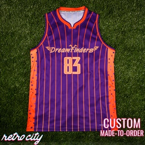 May include: Purple and orange basketball jersey with vertical stripes and orange stars on the sides. The jersey has the text "DreamFinders" and the number "83" on the front.