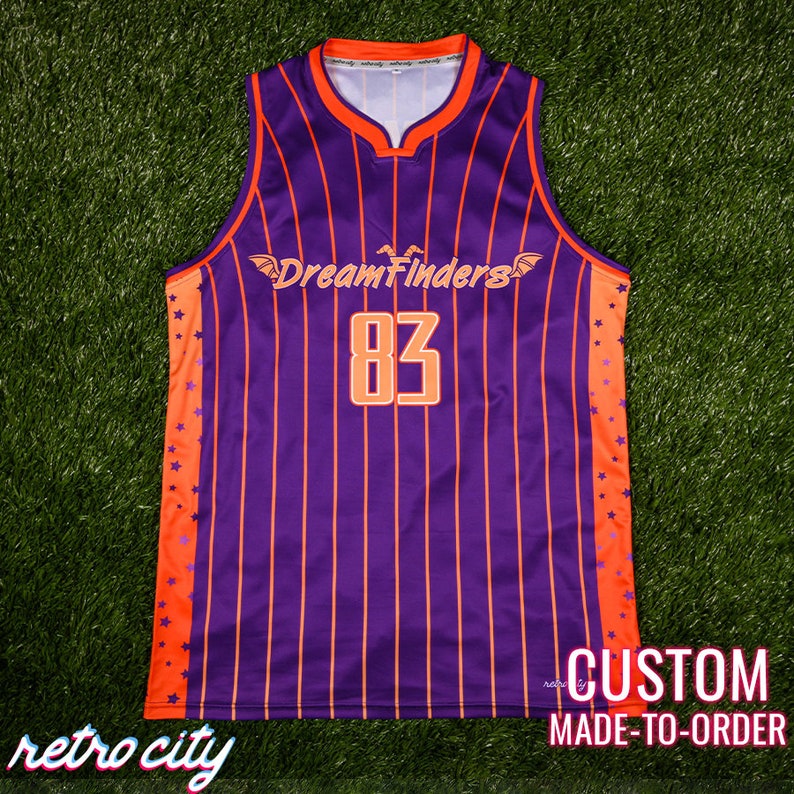 May include: Purple and orange basketball jersey with vertical stripes and orange stars on the sides. The jersey has the text "DreamFinders" and the number "83" on the front.
