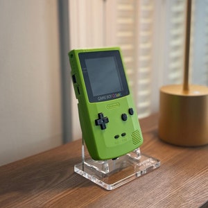 以下が含まれることがあります： ライムグリーンの任天堂ゲームボーイカラー携帯ゲーム機。黒い画面ベゼル、十字キー、2つの円形アクションボタンを備え、画面の下にはGame Boy Colorの文字がプリントされており、透明なアクリルスタンドに立てかけられています。