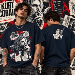 May include: A dark navy blue short-sleeve t-shirt featuring a graphic print of Kurt Cobain. The front displays a collage with his portrait, a guitar, and text including "Kurt Cobain," "the sun is gone, but i have a light," and "Peace, Love, Empathy 1967-1994."