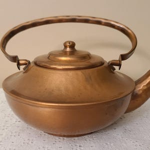 May include: A copper teapot with a hammered finish. The teapot has a rounded body, a curved handle, and a spout. The lid is topped with a small knob. The teapot is sitting on a white lace tablecloth. The teapot is approximately 10 inches wide.