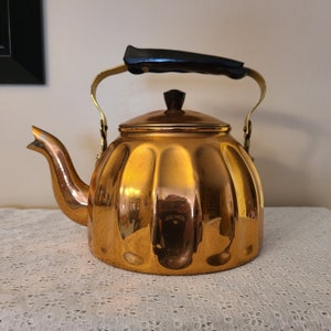 May include: A gold-colored copper kettle with a black handle and a fluted design. The kettle has a shiny finish and is sitting on a white lace tablecloth.