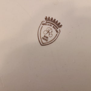 Puede incluir: Una marca de porcelana marr&oacute;n y blanca con el texto "Porcelaine Veritable" y "Revol France" dentro de una forma de escudo con una corona en la parte superior.