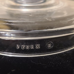 May include: A clear, circular glass lid featuring a stepped, concentric ring design on the top surface. The side of the lid is embossed with the text PYREX and a circled R symbol, indicating the brand and a registered trademark for this kitchenware.