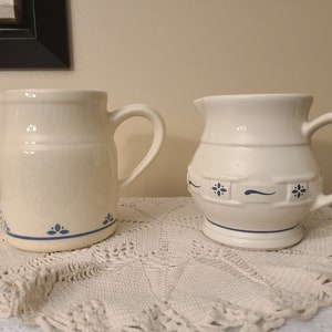 May include: Two white ceramic pitchers with blue floral designs. The pitcher on the left has a simple blue floral design around the bottom. The pitcher on the right has a more elaborate blue floral design around the middle.