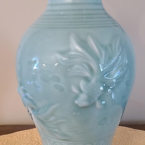 May include: A light blue ceramic vase with a flared rim and a narrow neck featuring horizontal ridges. The body of the vase displays a raised relief design of swimming fish and aquatic plants, while the base is decorated with vertical fluted lines.