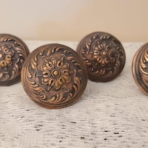 May include: Four round metal cabinet knobs with a dark bronze finish. Each knob features a raised central floral motif surrounded by intricate swirling leaf patterns. The surface of the knobs has a textured, weathered appearance with highlights.