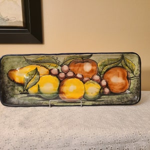 May include: A ceramic serving tray with a hand-painted design of fruit. The tray features a green, yellow, and orange color scheme with a variety of fruits, including apples, pears, and grapes.