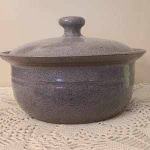 May include: A round, speckled lavender-gray ceramic pot with a matching lid. The pot features a wide, flared rim and a central knob on the lid. The surface has a textured, mottled appearance with subtle horizontal grooves circling the body.