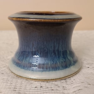 May include: A small, flared ceramic vessel featuring a deep blue and brown reactive glaze. The exterior displays a vertical drip pattern that transitions from a dark brown rim to a lighter blue body, ending in a solid white base with a smooth finish.
