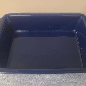 May include: A rectangular navy blue ceramic baking dish with a glossy finish and integrated handles on the short sides. The dish features a deep basin with smooth, rounded interior corners and a wide, flat rim extending around the entire perimeter.