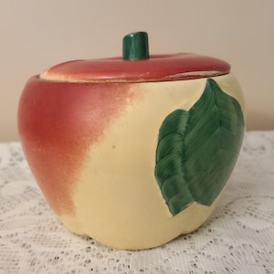 May include: A ceramic apple-shaped jar with a lid. The jar is painted with a gradient of red and cream colors, and a green leaf design. The lid has a green stem. The jar is likely used for storage.