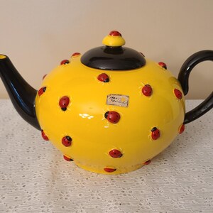 May include: A yellow ceramic teapot with a glossy finish, adorned with red ladybug accents. The teapot features a black handle, spout, and lid, creating a striking contrast. A small label is affixed to the side. A charming addition to any kitchen.
