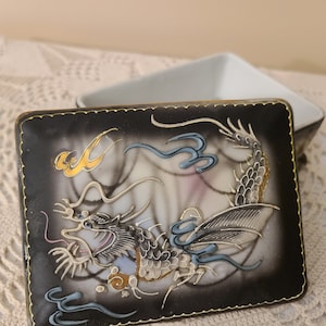May include: A black and white porcelain trinket box with a gold trim. The lid features a detailed painting of a dragon with blue and gold accents.