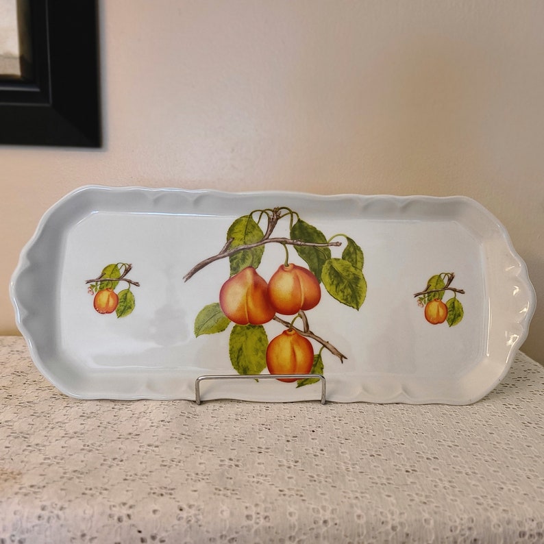 Puede incluir: Bandeja de servir de cer&aacute;mica blanca con un dise&ntilde;o decorativo de una rama de fruta. La fruta se representa en tonos de naranja y rojo. La bandeja tiene un borde festoneado.