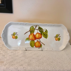 Puede incluir: Una bandeja de servir de cer&aacute;mica blanca con borde festoneado presenta un dise&ntilde;o pintado a mano de una rama con fruta naranja y hojas verdes.