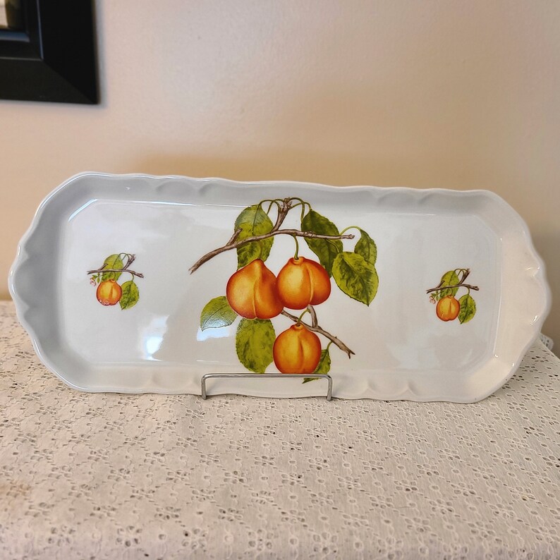 Puede incluir: Una bandeja de servir de cer&aacute;mica blanca con borde festoneado presenta un dise&ntilde;o pintado a mano de una rama con fruta naranja y hojas verdes.