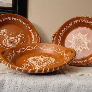 May include: Three terracotta ceramic plates with white painted designs. The plates have a raised rim and a handle on each side. The designs include a stylized bird, a flower, and a heart.