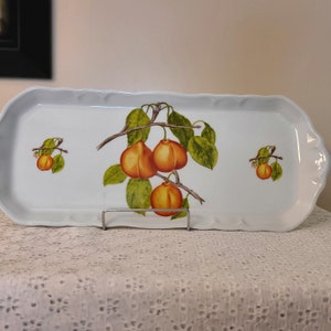 Puede incluir: Una bandeja de servir de cer&aacute;mica blanca con borde festoneado presenta un dise&ntilde;o pintado a mano de una rama con albaricoques naranjas y hojas verdes.