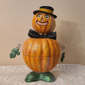 May include: A whimsical Halloween pumpkin figure with a smiling face, wearing a black bow tie and hat with a yellow band. The pumpkin has a green glittery base and decorative metal vines.