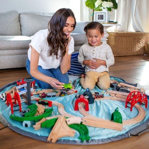Puede incluir: Un juego de tren de juguete de madera con una alfombra de juego azul. El juego incluye v&iacute;as de madera, puentes, t&uacute;neles y varios vagones. Una mujer y un ni&ntilde;o juegan con el tren en el suelo.