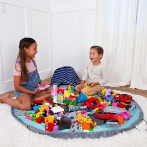Puede incluir: Una alfombra de juego azul claro y gris con borde de cord&oacute;n, llena de bloques de construcci&oacute;n coloridos. Dos ni&ntilde;os est&aacute;n sentados en la alfombra, jugando con los bloques. Una bolsa a rayas azules y blancas tambi&eacute;n est&aacute; en la alfombra.