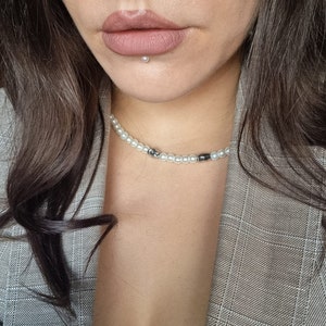 May include: A woman wearing a gray plaid blazer with a pearl and black bead necklace. She has a small pearl stud in her lower lip.
