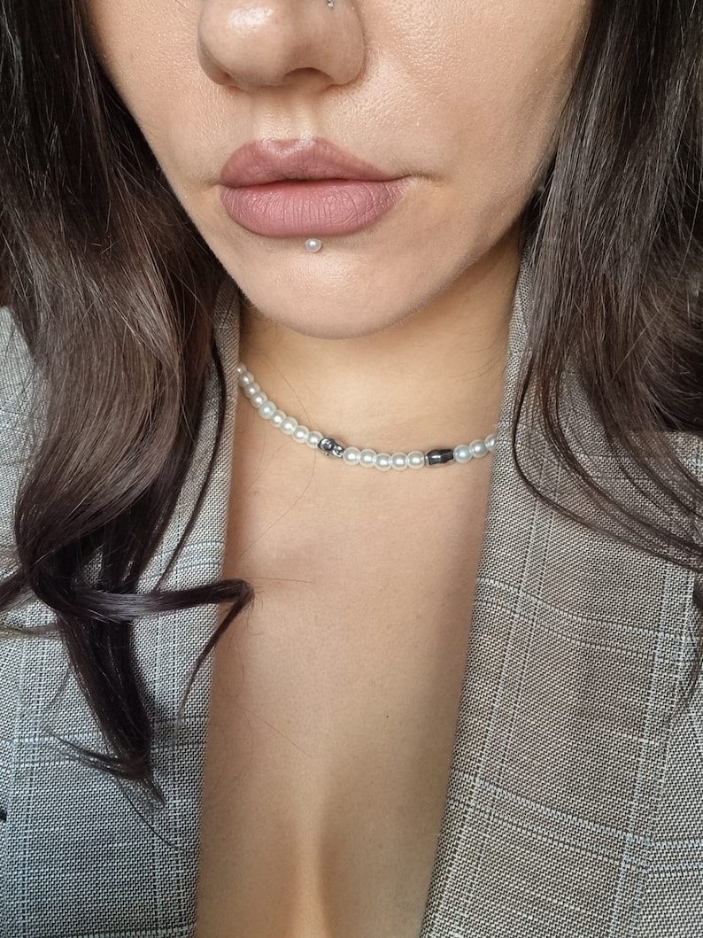 May include: A woman wearing a gray plaid blazer with a pearl and black bead necklace. She has a small pearl stud in her lower lip.